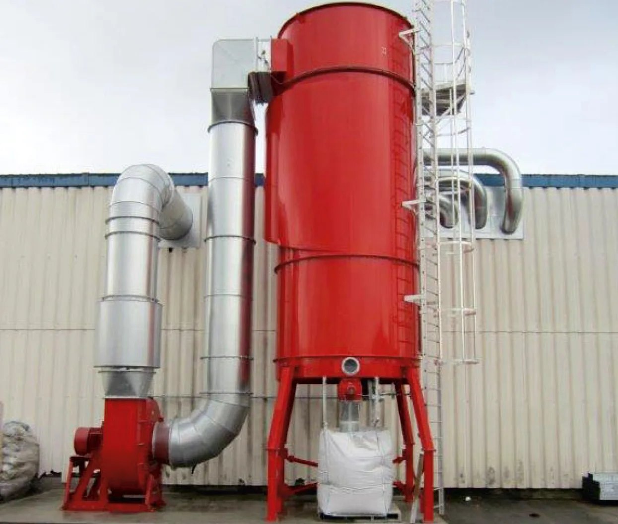 Un dépoussiéreur industriel rouge avec un réseau de gaines raccordé et un sac de collecte à l’extérieur d’une usine.